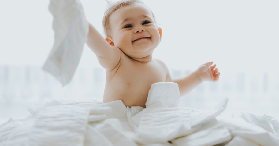 baby holding white disposable diapers sitting in huge pile of diapers