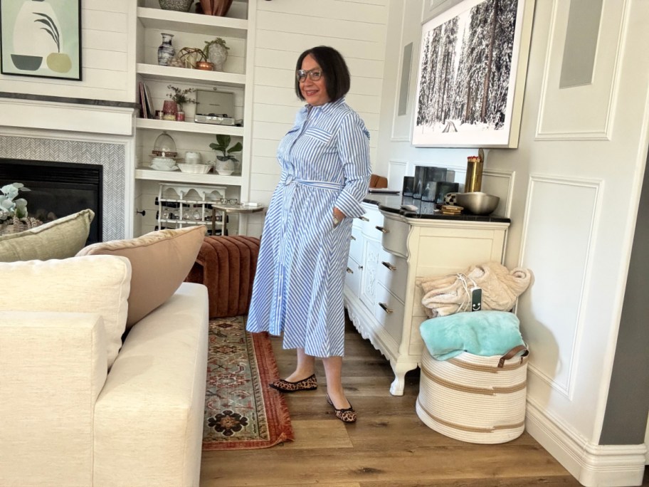 smiling woman standing in a living room, wearing a blue and white striped belted dress