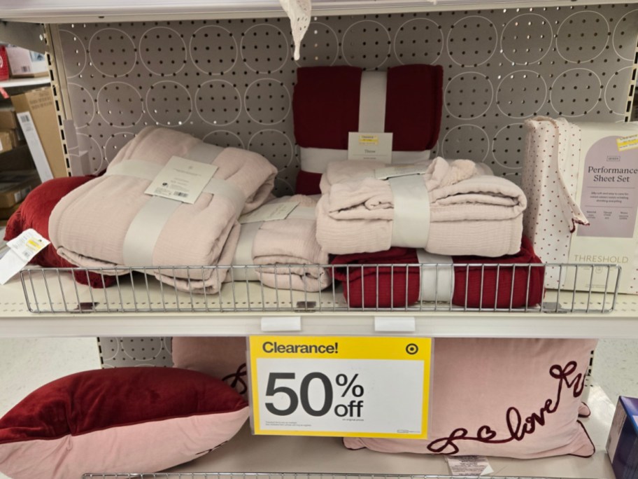 Clearance sale display of pink and red bedding, including folded blankets and pillows, on a shelf. Sign indicates 50% off.