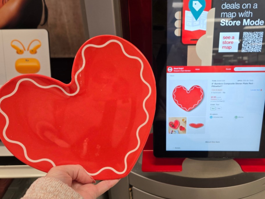 A red, heart-shaped plate with a wavy white border is held in front of a touchscreen showing the same plate for sale.