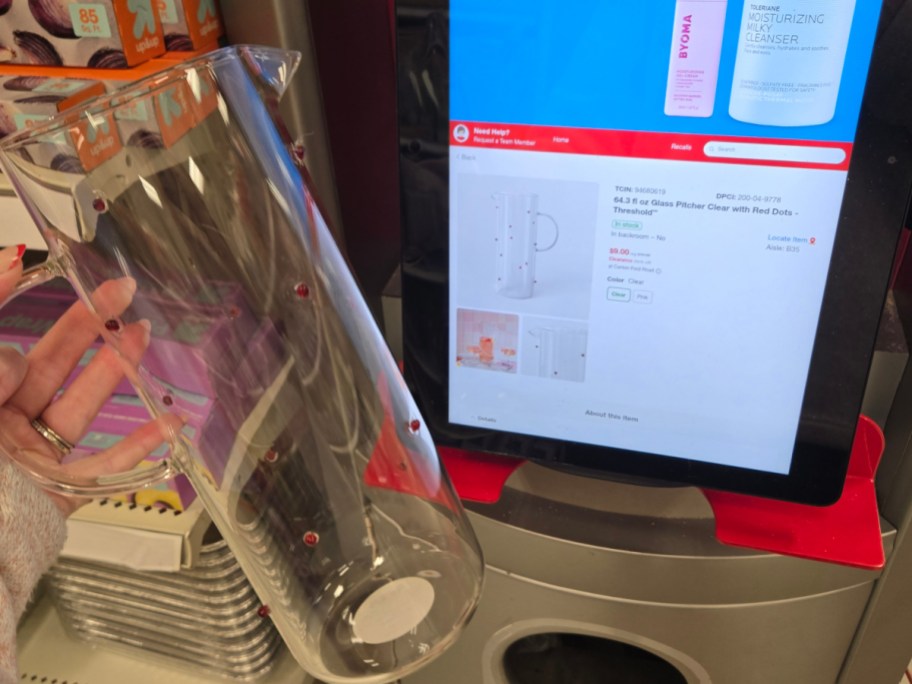 A person holds a clear glass pitcher with red dots next to a tablet screen displaying the same item for sale online.