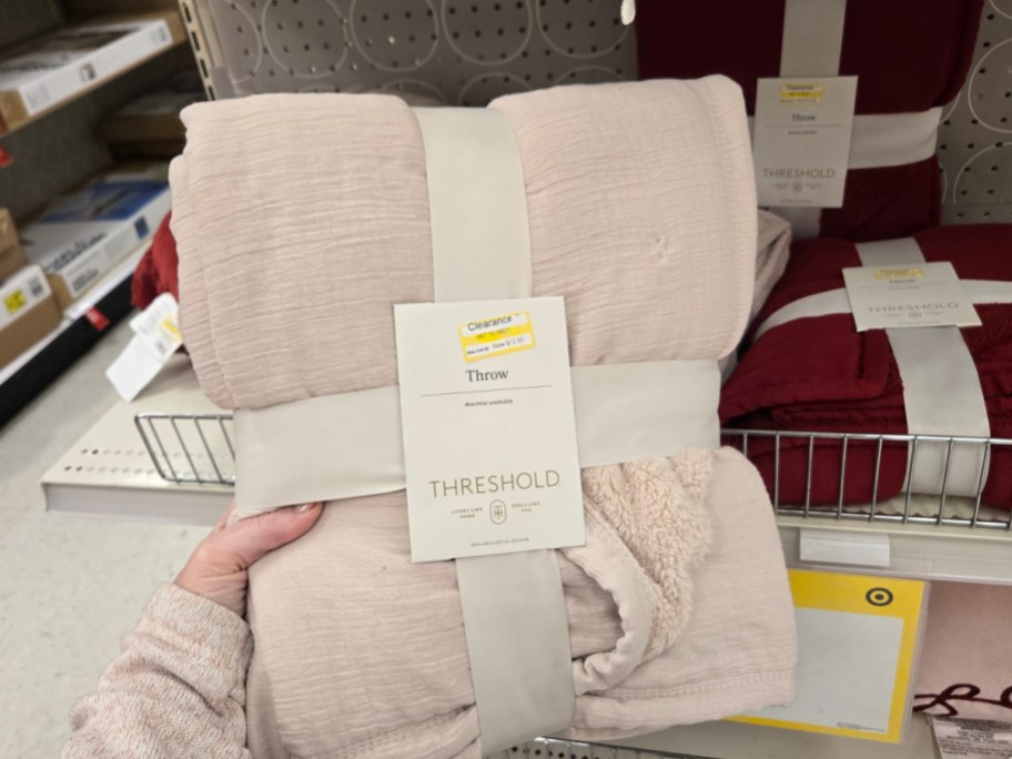 A pink Threshold throw blanket is displayed on a store shelf with a clearance label. Shelving and red blankets are visible in the background.