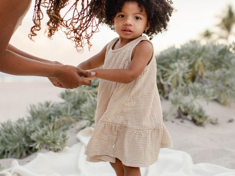 Burts bees baby girl wearing a tan dress on the beach holding her mothers hand