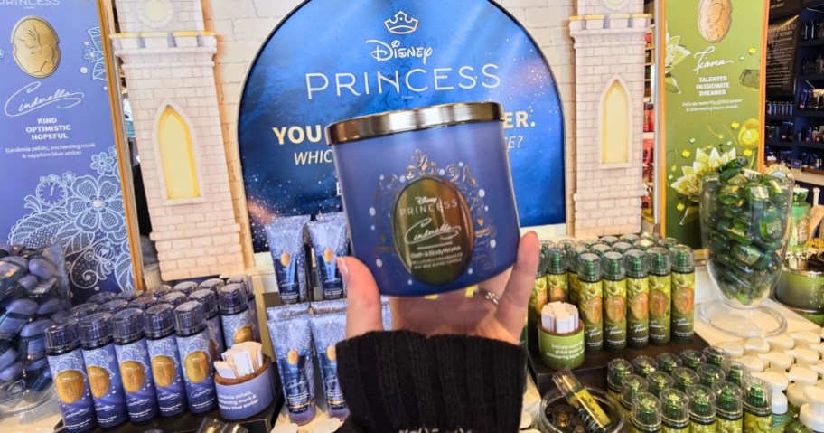 hand holding a blue Disney Princess Cinderella Candle up in a Bath & Body Works store, Disney Princess Collection display is behind it
