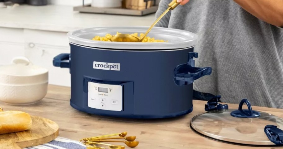 a blue Crock Pot on a kitchen counter with a person stirring the food inside it