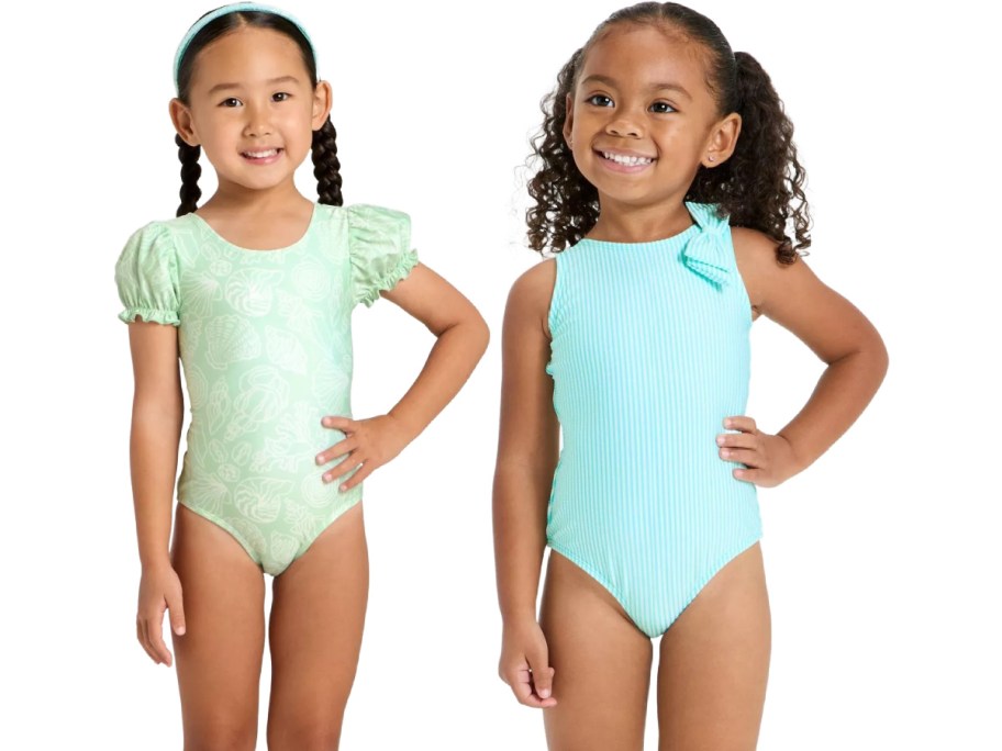 Two young girls smiling in swimsuits. The left girl wears a light green suit with puffed sleeves; the right girl wears a light blue sleeveless suit.