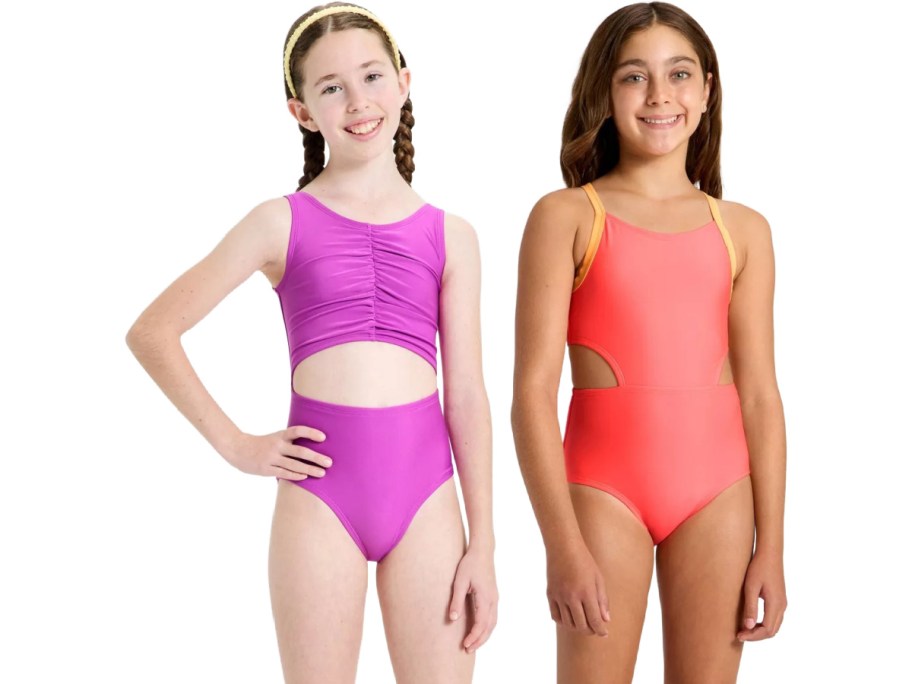 Two girls wearing vibrant swimsuits stand smiling. The left girl wears a purple suit with cut-out detail; the right girl wears a coral suit with yellow straps.