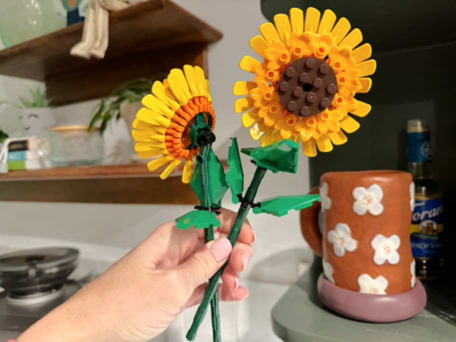 hand holding two assembled lego sunflowers in front of shelves and a coffee mug