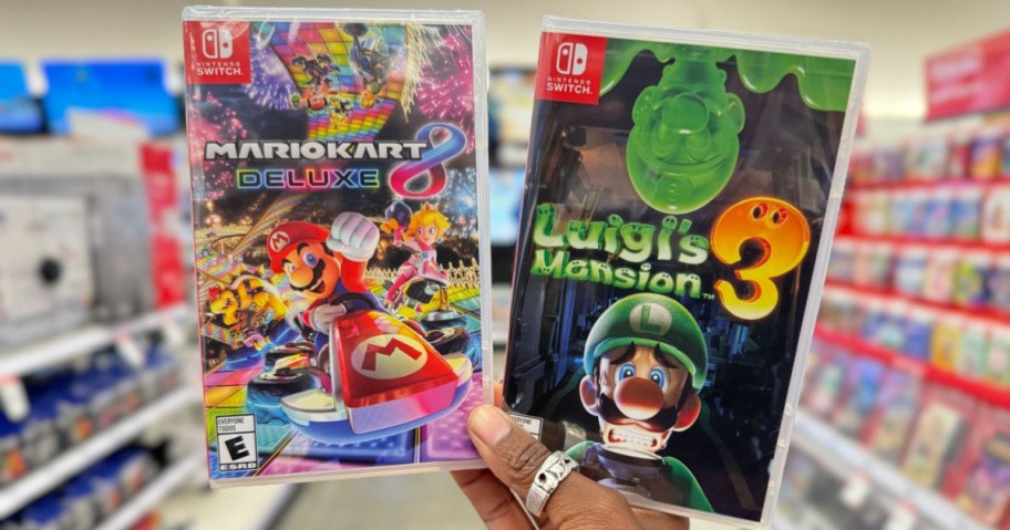 Close-up of hands holding two Nintendo Switch game cases in a store. Left: "Mario Kart 8 Deluxe" with colorful racing scene. Right: "Luigi's Mansion 3" featuring Luigi looking scared.