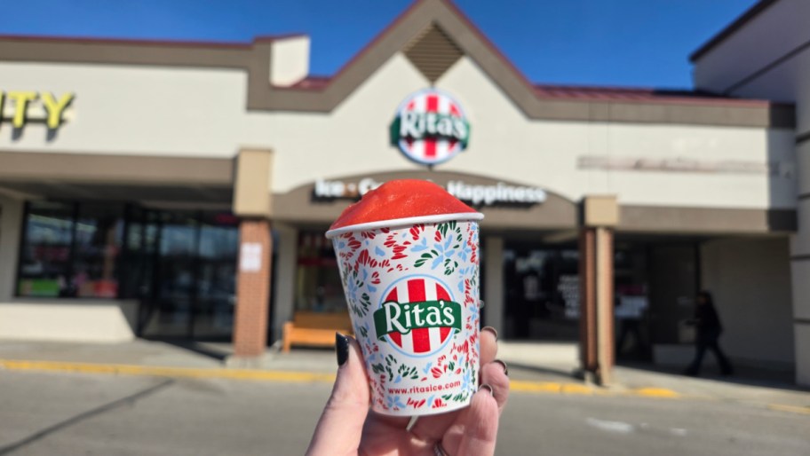 hand holding up an italian ice in a cup with a rita's restaurant in the background