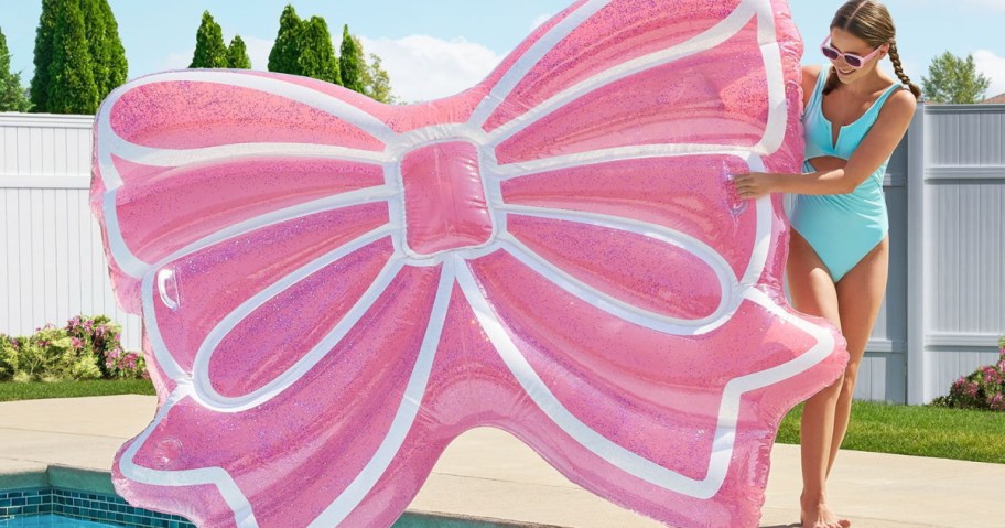 A woman in a turquoise swimsuit and sunglasses holds a large pink bow-shaped pool float at the edge of a pool on a sunny day.