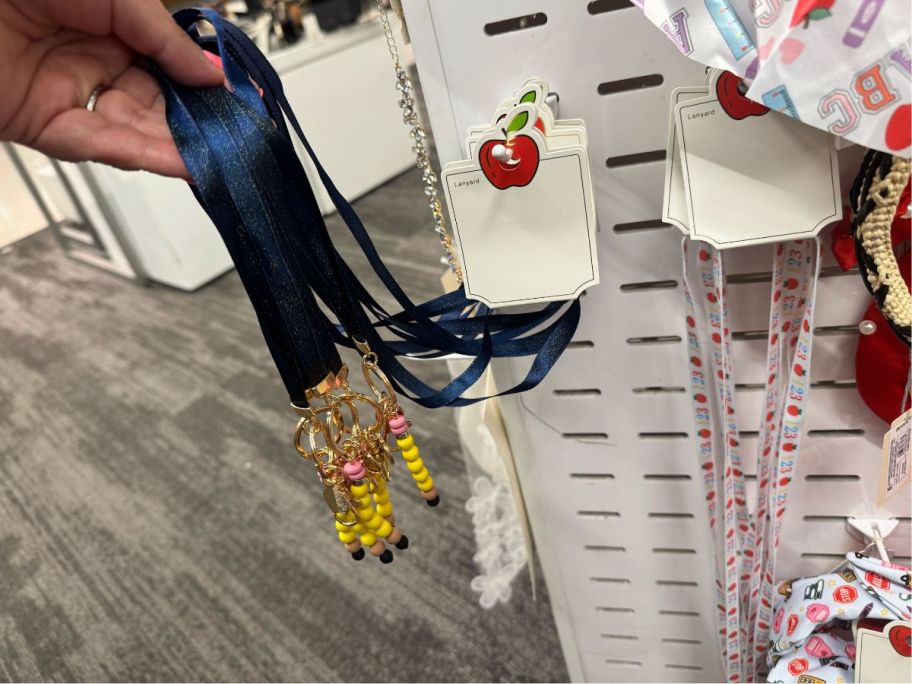 Teacher Pencil & Apple Charm Sparkle lanyard in hand in store and Teacher Apple & 123 Print w/ Charms Lanyard hanging next to it 