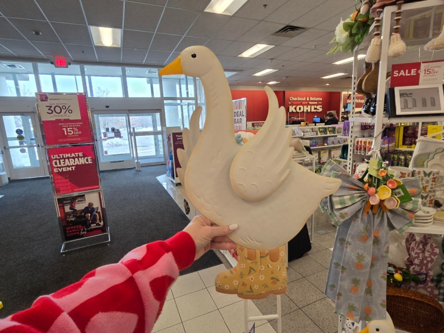 Person holding a decorative duck wearing rain boots next to a bow in a Kohl's store entrance with sale signs and promotional displays visible.