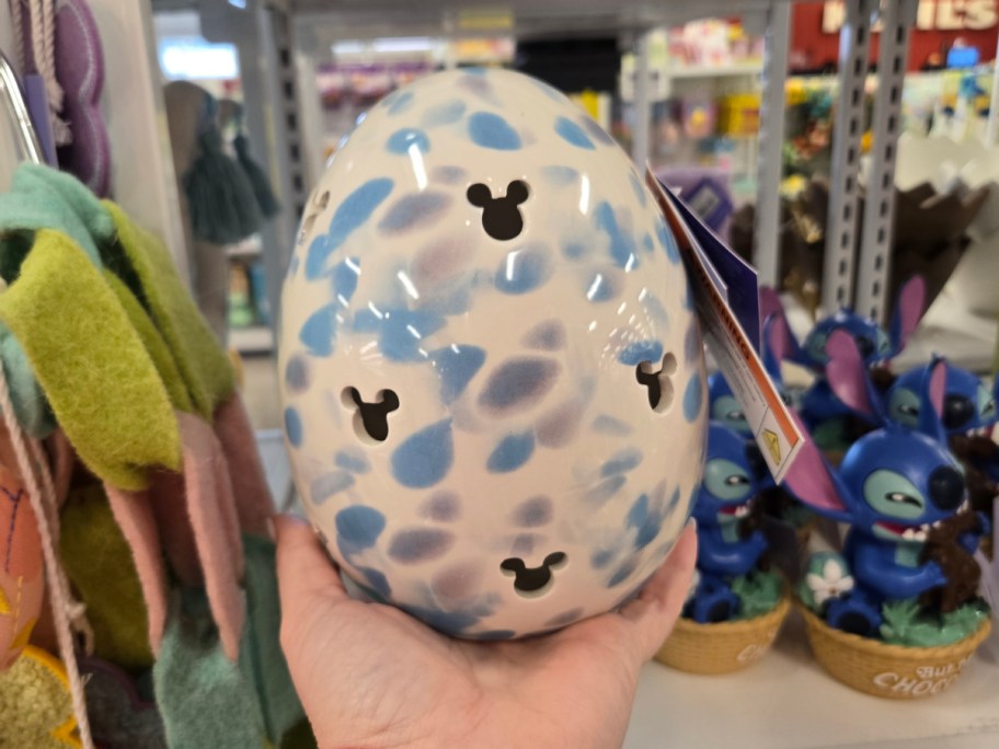 Hand holding a speckled ceramic egg adorned with black Mickey mouse-shaped cutouts, featuring blue and purple spots, situated in a store with themed merchandise.
