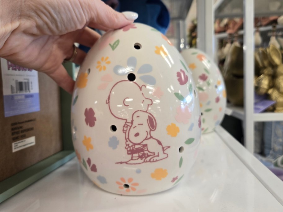 A hand holding a decorative ceramic egg with pastel flowers and a peanuts Snoopy and Charlie Brown illustration, featuring small holes. 