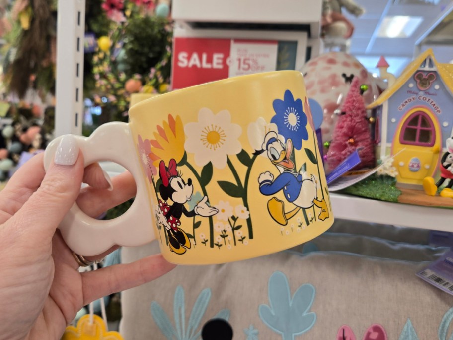 Yellow mug featuring Minnie and Donald Duck among colorful flowers. Displayed in a store with sale signs.