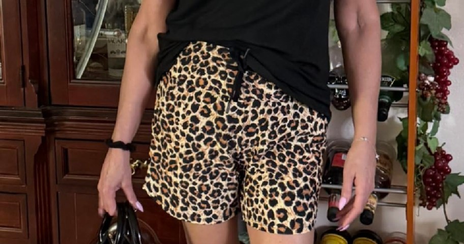 A person stands indoors wearing leopard-print shorts and a black top, holding a handbag. They are near a wine rack and wooden cabinets.