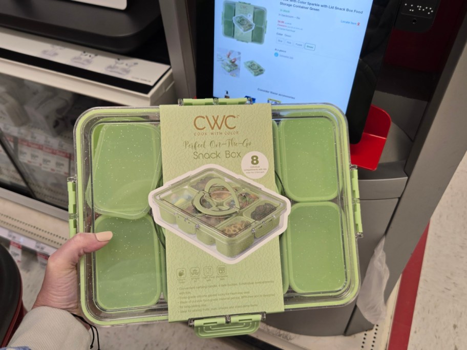 A hand holds a green snack box set with eight compartments in front of a store display. The box is displayed alongside an online listing on a screen.