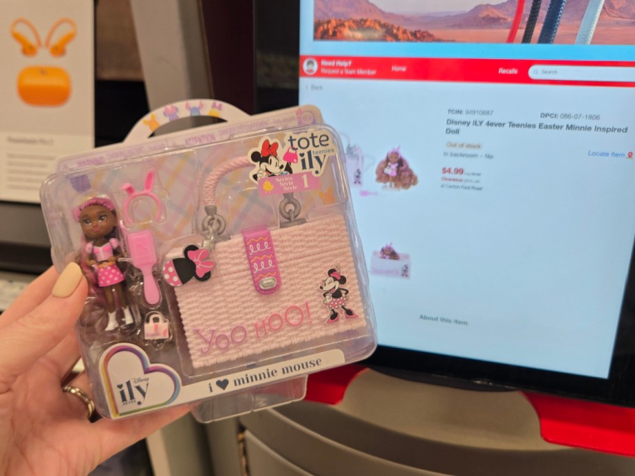 A hand holds a Disney ILY 4EVER Minnie Mouse doll set, featuring a small doll, accessories, and a tote bag. In the background, a store price scanner displays the item details.