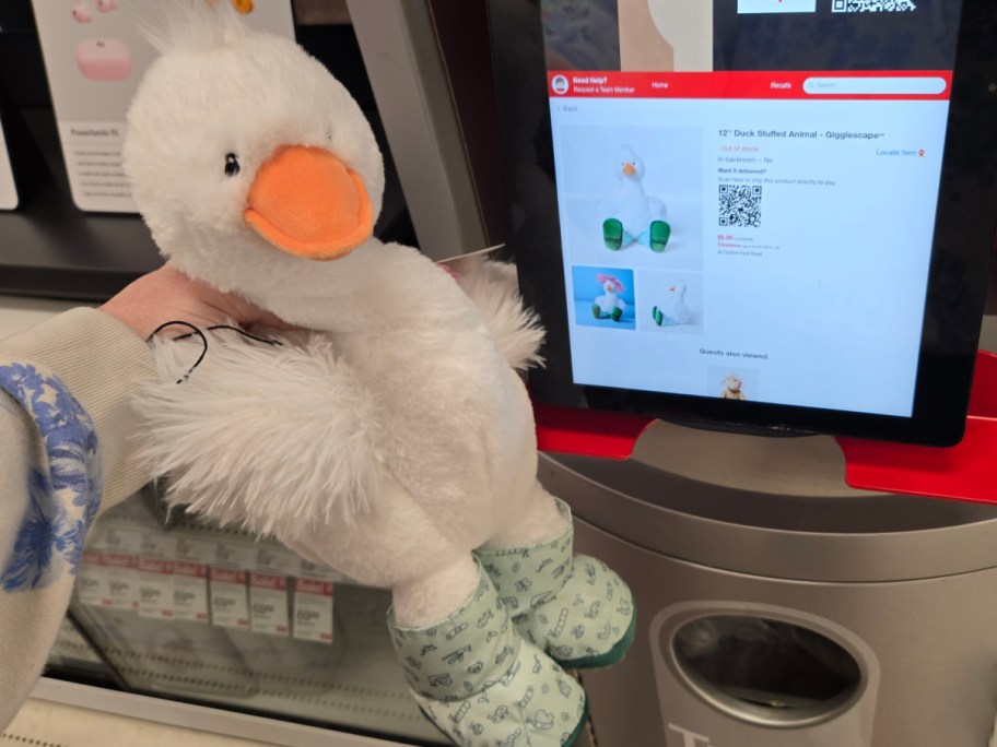 A hand holding a fluffy white duck stuffed animal with an orange beak, facing a store scanner displaying its product details on the screen.