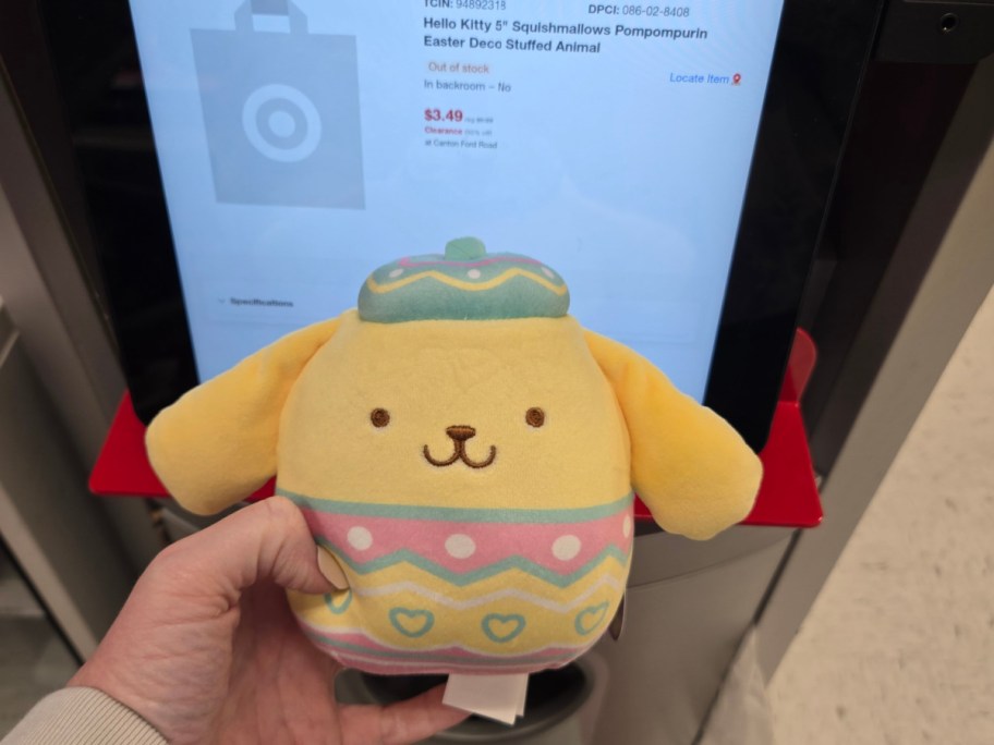 A hand holds a yellow Pompompurin stuffed toy with pastel patterns in front of a store's price scanner display.