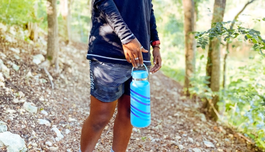 person standing on hiking trail in woods holding blue water bottle from handle