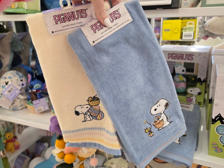 Two hand towels featuring Peanuts characters are displayed. One is cream-colored with Snoopy carrying Easter eggs, and the other is blue with Snoopy holding a basket.