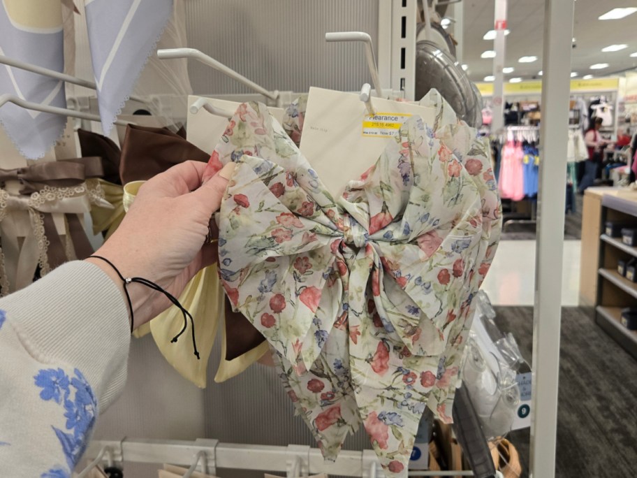 A hand holds a floral-patterned bow on clearance in a store aisle, with various fabrics and clothing items visible in the background.