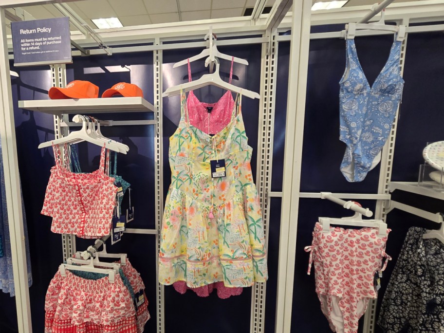 Retail display of colorful clothing and accessories, featuring a floral dress, patterned swimsuits, and orange hats. A return policy sign is visible.