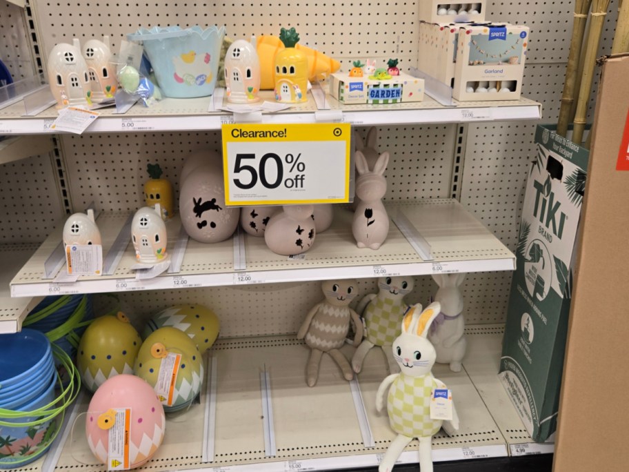 Store shelf with decorative items on clearance, featuring bunny and carrot figurines, pastel Easter eggs, and fabric toys. "50% off" sign promotes discounts.