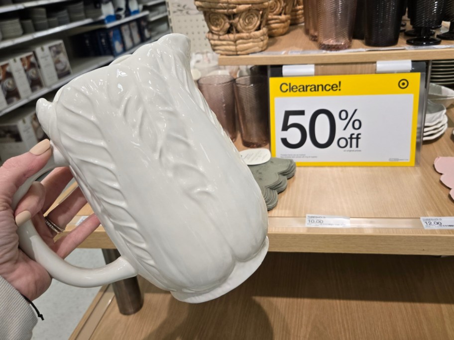 Person holding a white, floral-patterned ceramic pitcher in a store. A yellow clearance sign shows "50% off" on shelves with various home goods.