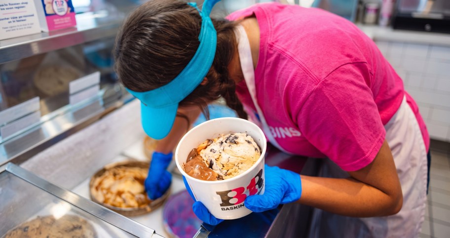 A Baskin-Robbins employee wearing a blue visor, pink t-shirt, and blue gloves leaning over a counter while scooping ice cream into a Baskin-Robbins paper cup.