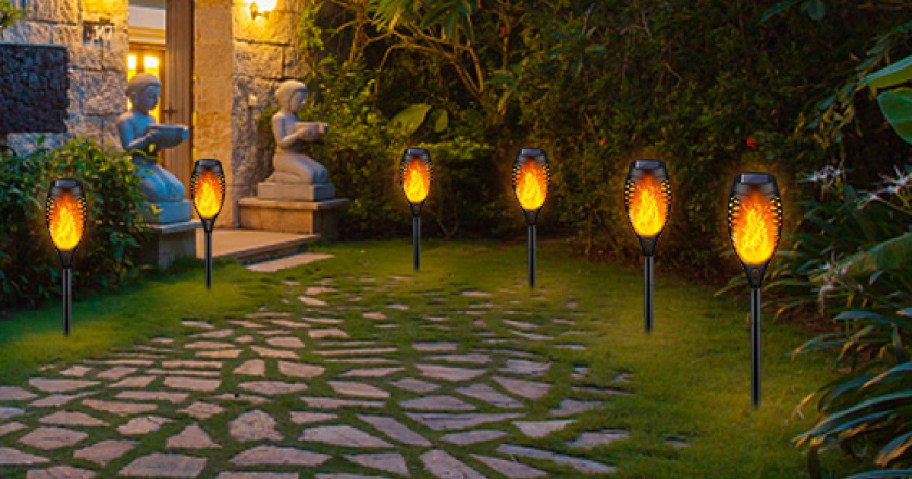 Stone pathway illuminated by glowing torches leads to a house entrance. Two stone statues sit near the door, surrounded by lush greenery.