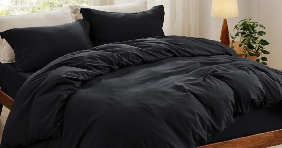 A cozy bed features a plush black duvet and pillows with crisp white sheets. A wooden nightstand holds a lit lamp and a leafy plant.