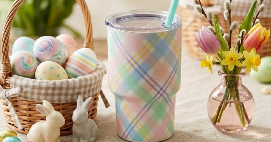 Plaid tumbler with pastel colors, a blue straw, sits beside a basket of decorated Easter eggs. Ceramic bunnies and spring flowers.