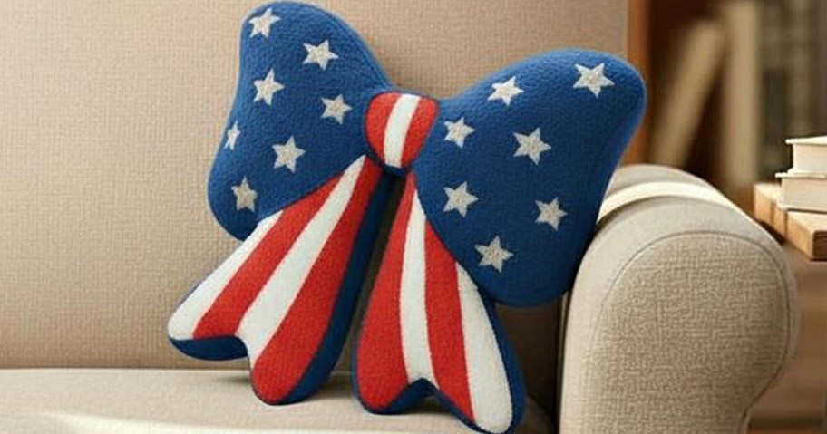 A plush bow pillow on a beige couch, patterned with American flag colors; blue with white stars and red and white stripes.