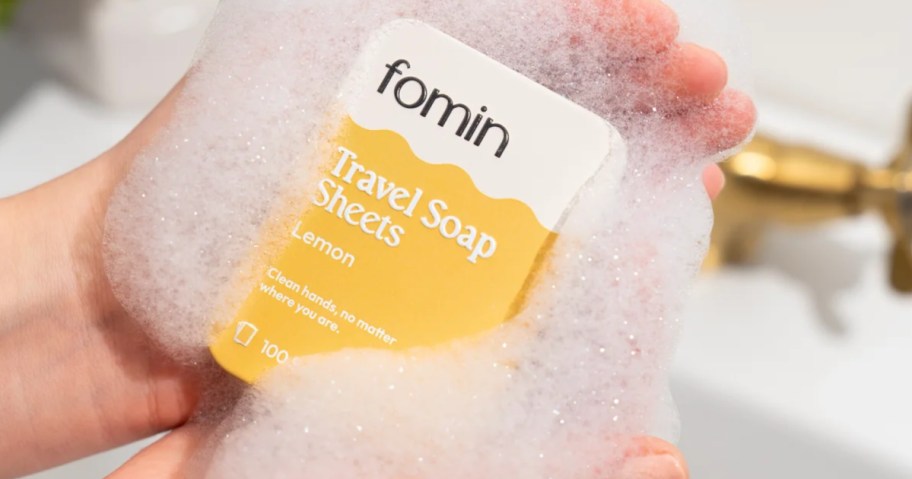 Person holding yellow travel sized box of soap, sheets over sink