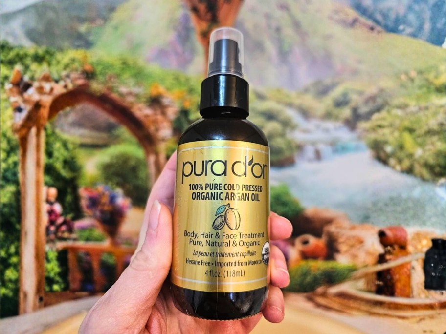 hand holding a bottle of Pura D'or Organic Moroccan Argan Oil in front of a wall mural