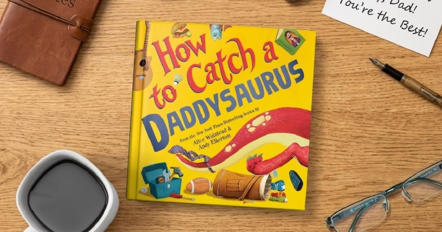 A colorful children's book titled "How to Catch a Daddysaurus" lies on a wooden table, surrounded by a coffee cup, glasses, and a handwritten note.