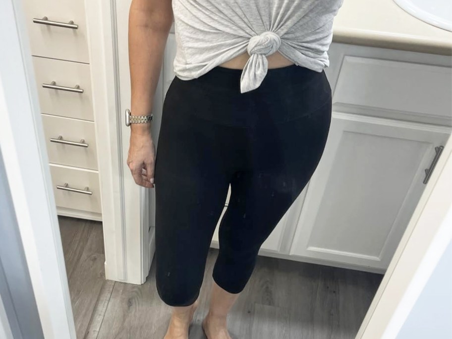 woman taking photo in mirror wearing light grey top and black capri leggigns