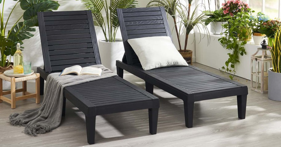 Two dark wooden lounge chairs with white cushions and a gray blanket, set in a bright room with plants and a small table holding lemonade.