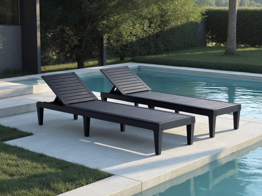 Two sleek black lounge chairs sit poolside on a light stone patio, surrounded by lush greenery.