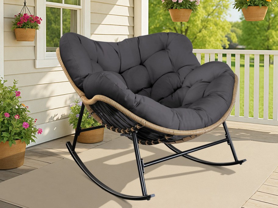 A cozy, black cushioned rocking chair sits on a porch, surrounded by hanging and potted flowers.