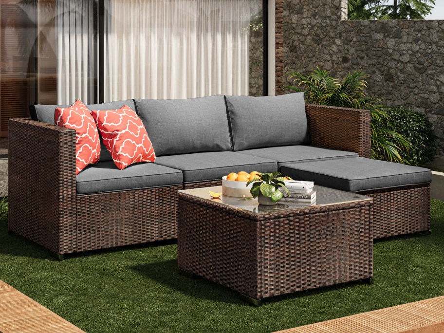 Outdoor patio with a wicker sectional sofa featuring gray cushions and red patterned pillows. A matching wicker table holds a bowl of oranges and books.