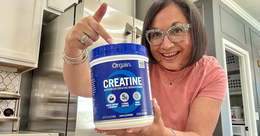 Woman in Glasses holding and pointing to a Orgin Creatine Jar while smiling