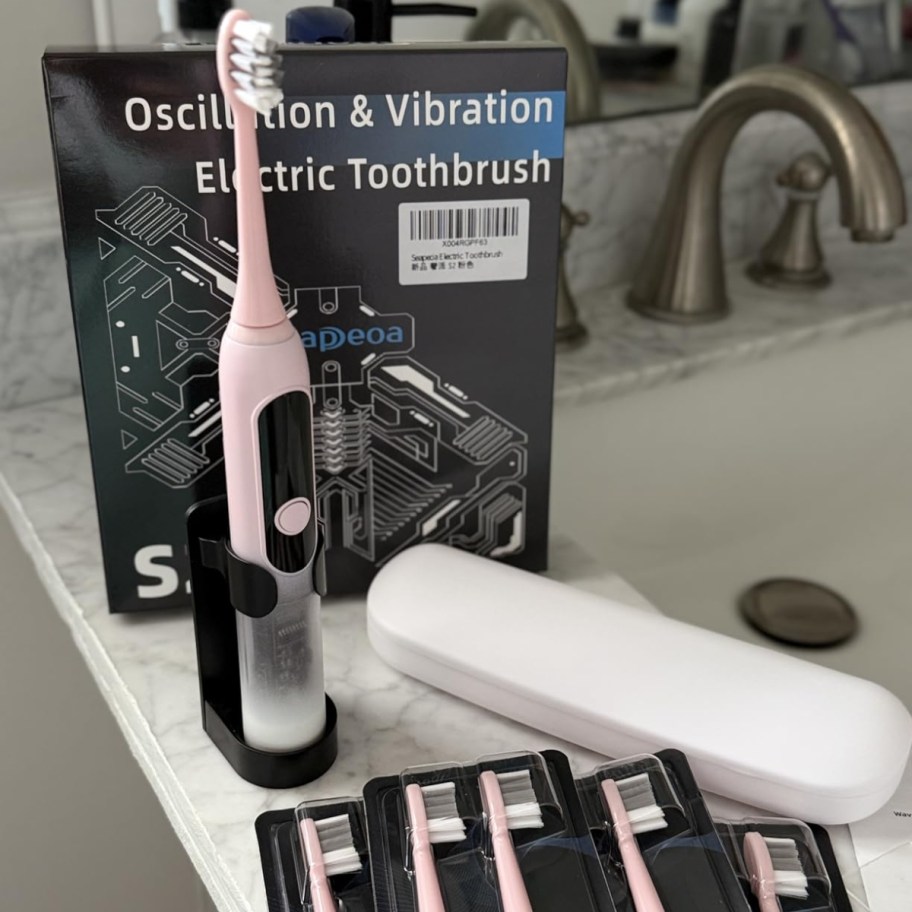 a pink electric toothbrush in a stand with extra heads and a white case around it, the box it comes in behind it