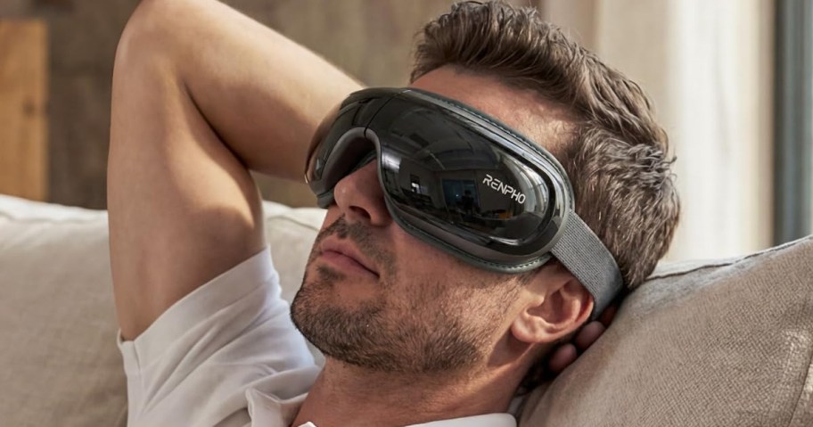 A man relaxes on a beige couch wearing a high-tech eye massager with a dark visor.