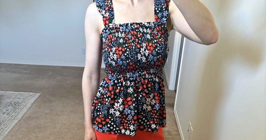 Woman in a floral sleeveless top with red and white flowers takes a selfie in a room with beige carpet.