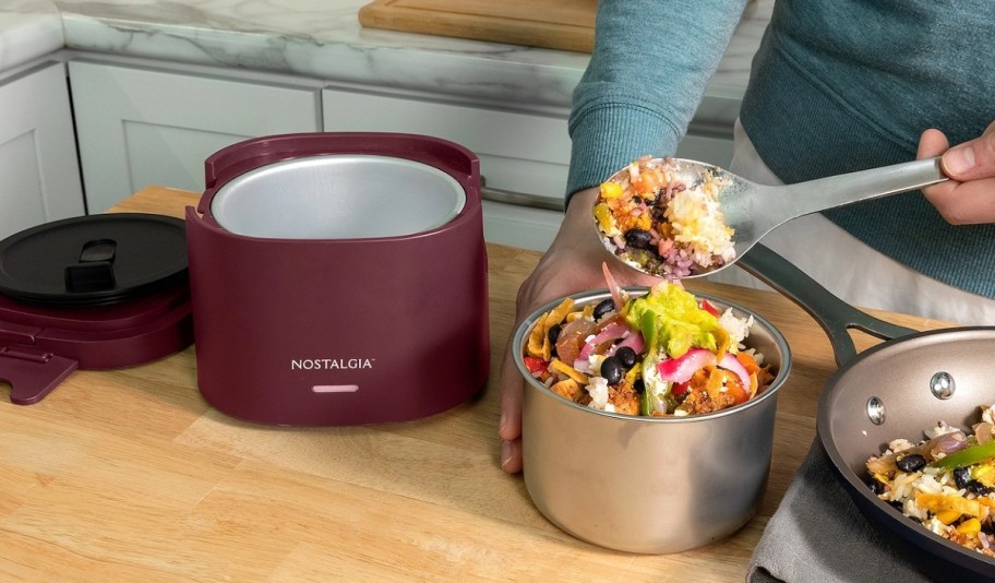 a burgundy electric lunch box on a kitchen counter as a woman places food in it
