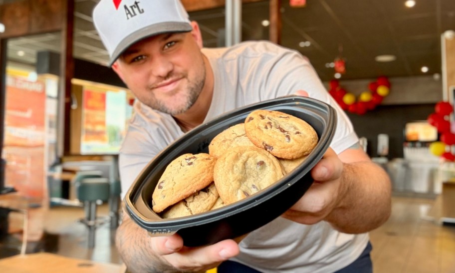 a man in a cap holding a black palstic bowl filled with chocolate chip cookies